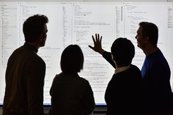 Four people, who can only be recognised in outline, look at a projection of software code on a screen.