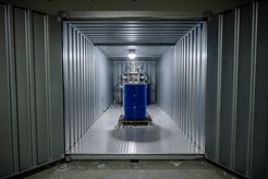 Blick in einen hell erleuchteten Container aus Wellblech, im Zentrum steht ein blaues Fass, umgeben von einer Konstruktion.