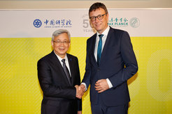Two men in business suits are shaking hands. The man on the left, with short, graying hair, is wearing a dark navy or black suit, a light-colored collared shirt, and a dark tie. He is middle-aged, and appears to be of East Asian descent, judging by his facial features. He is smiling and looking directly at the camera. The man on the right, with short brown hair and glasses, is also wearing a dark navy or black suit, a light colored collared shirt, and a teal-blue tie. He is middle-aged and appears to be of European descent. He is also smiling and looking directly ahead, though his focus is slightly turned toward the camera. The background is a bright, mustard-yellow backdrop with a repeating patterned design that resembles a grid or woven texture. Logos or banners, containing Chinese and English text, are visible on the wall behind the men. The banners seem to be related to science or an academic institution. The typography and graphic elements suggest a celebratory event or an anniversary. The 1973-2023 or similar numbers and anniversary wording are on the backdrop.