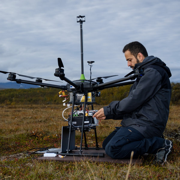 A man with black clothes is sitting in a landscape and working on an uncrewed aerial vehicle with scientific equipment
