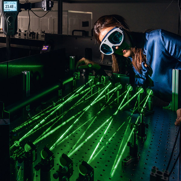 A woman wearing a blue blouse and safety goggles looks at an optical table with green laser beams.