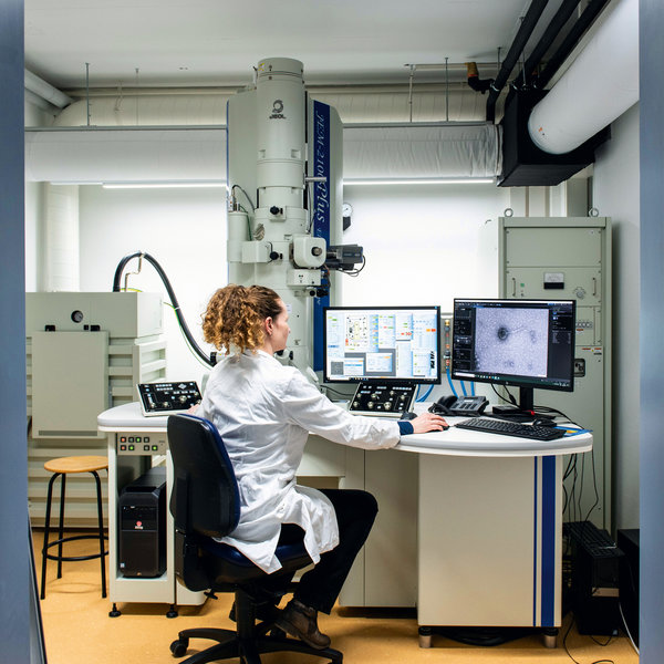 A woman in a white coat sits in front of a large microscope and a computer