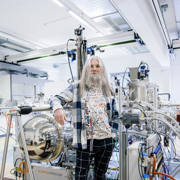 A person wearing colourful clothing is standing in a laboratory with lots of equipment, tubes and cables